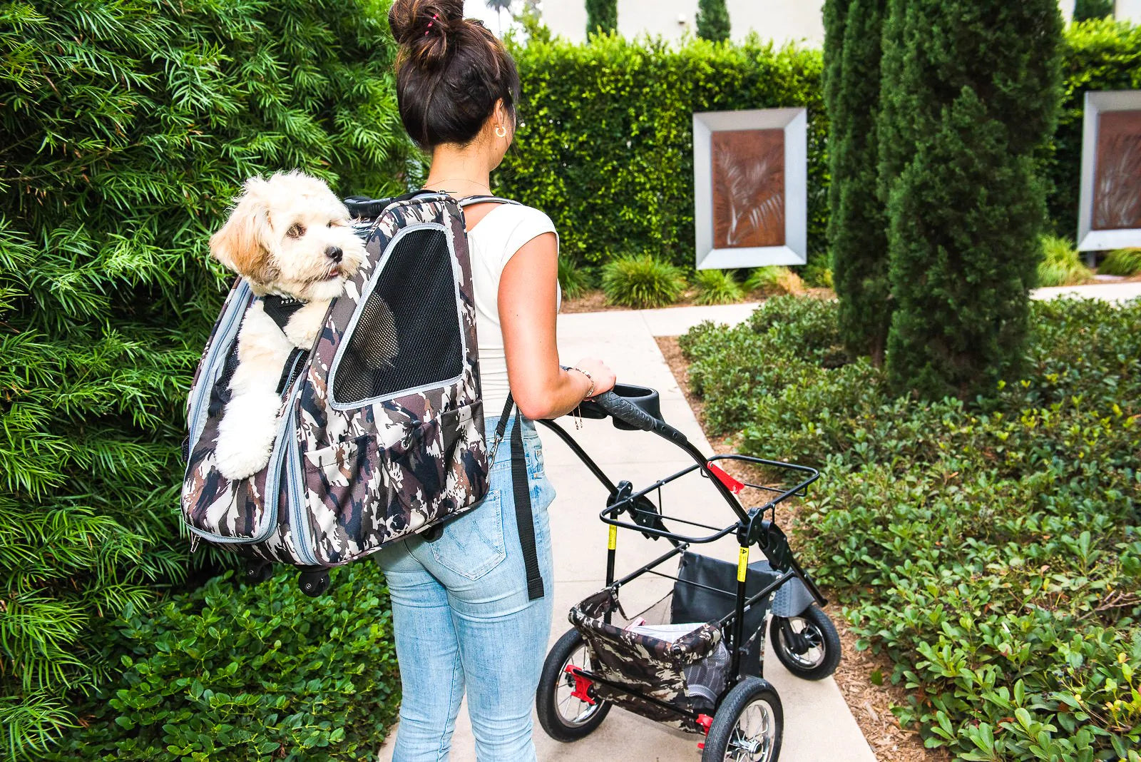 A woman pushing a Petique dog stroller frame carrying a small dog inside a Petique pet carrier backpack.